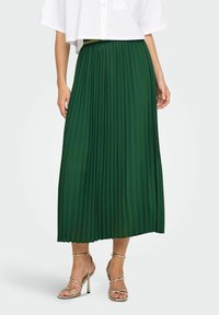 Gonna maxi plissettata verde con vita elasticizzata e tessuto fluido, abbinata a sandali con tacco alto e cinturini metallici.