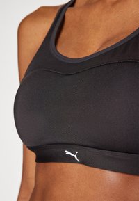 Svart sportsbh laget av elastisk stoff, med en scoop-halsringning og en hvit Puma-logo på båndet. Jevn tekstur med minimale sømmer.