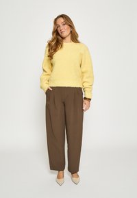 Sieviete ar gariem viļņainiem matiem, ģērbusies dzeltenā pūkainā džemperī, brūnās platās biksēs, bēšās garajās kurpēs un ar brūnu rotaslietu, stāv.