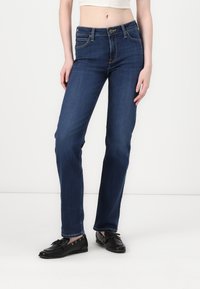 Jeans de mezclilla de un azul oscuro con una cintura ajustada, diseño de pierna recta, cinco bolsillos y costuras en contraste. Llevados con zapatos negros de tipo slip-on.
