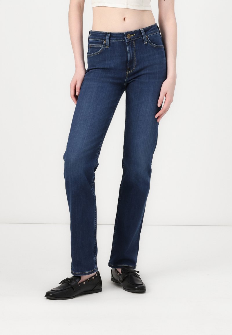 Jeans de mezclilla de un azul oscuro con una cintura ajustada, diseño de pierna recta, cinco bolsillos y costuras en contraste. Llevados con zapatos negros de tipo slip-on.