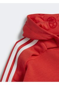 Felpa rossa realizzata in tessuto morbido, con un collo alto e strisce bianche lungo le maniche, con una stampa discreta del logo all'interno del cappuccio.