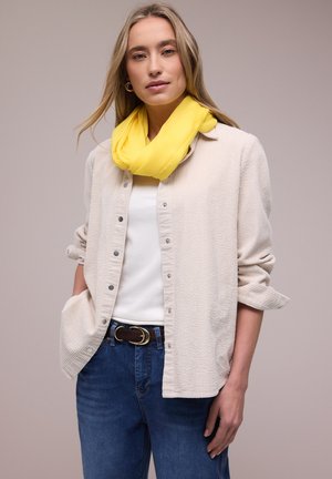 Frau mit gelbem Schal, cremefarbener, geknöpfter Bluse mit hochgekrempelten Ärmeln, weißem Top, blauen Jeans und schwarzem Gürtel vor einem neutralen Hintergrund.