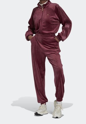 Persoon draagt een bordeauxrode velours trainingspak met een jas met rits en broek met elastische boorden, gecombineerd met witte chunky sneakers.