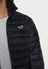 Black puffer jacket with a high collar and horizontal quilted design. Features a white logo on the chest and a smooth, shiny texture.