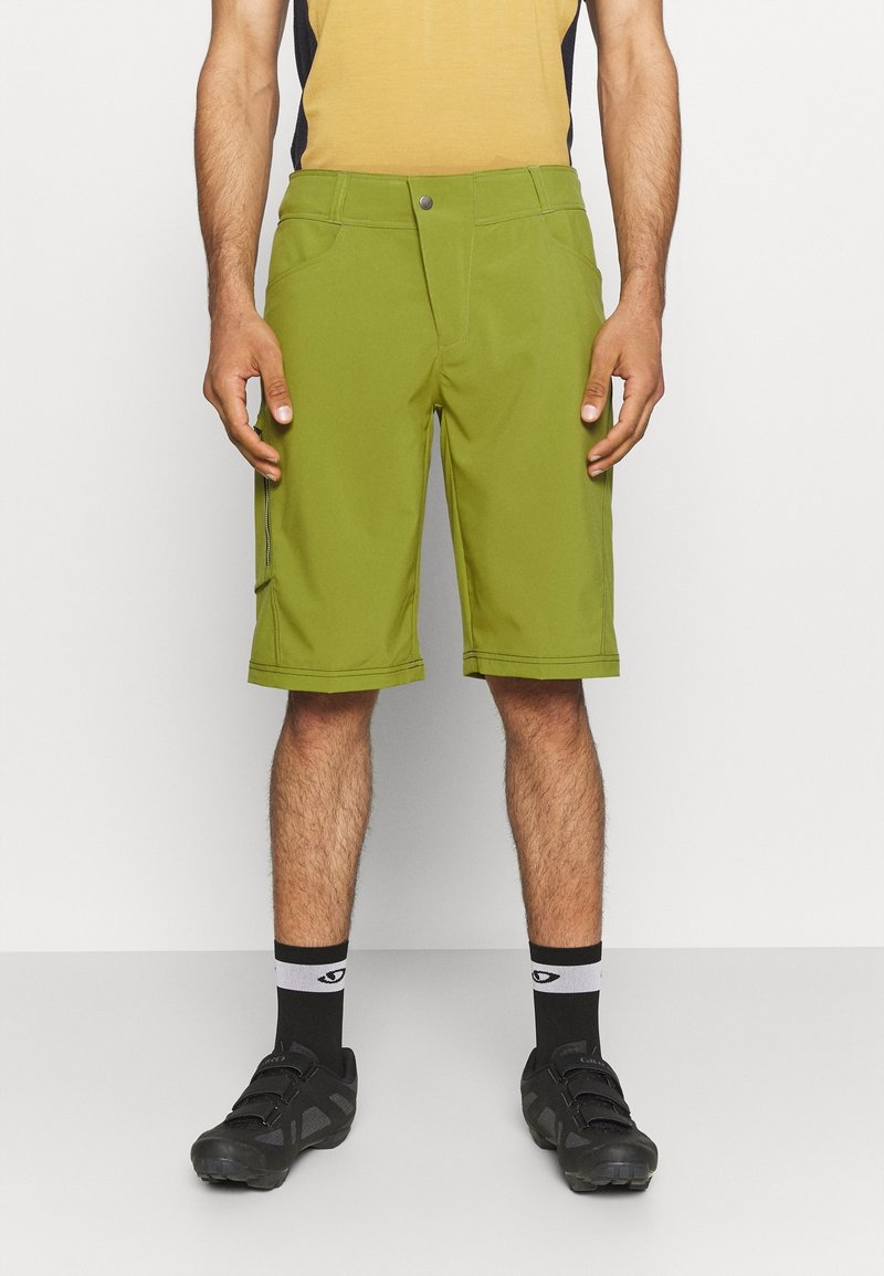 Homme portant un short vert olive longueur genou, des chaussettes noires et blanches et des chaussures de sport noires, debout devant un fond clair uni.