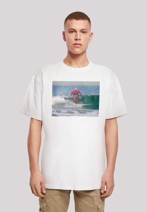 White oversized t-shirt featuring a colorful surfing graphic, depicting a surfer in a pink wetsuit riding a wave. Casual fit, short sleeves.