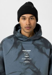 Blauwe tie-dye hoodie met een zachte textuur, voorzien van witte bedrukt tekst op de voorkant. Het model draagt een zwarte ribbelbeanie.