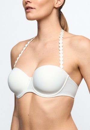 Vrouw draagt een witte strapless beha met geschulpte halterneck bandjes, getoond van schouders tot middenrif tegen een effen achtergrond.