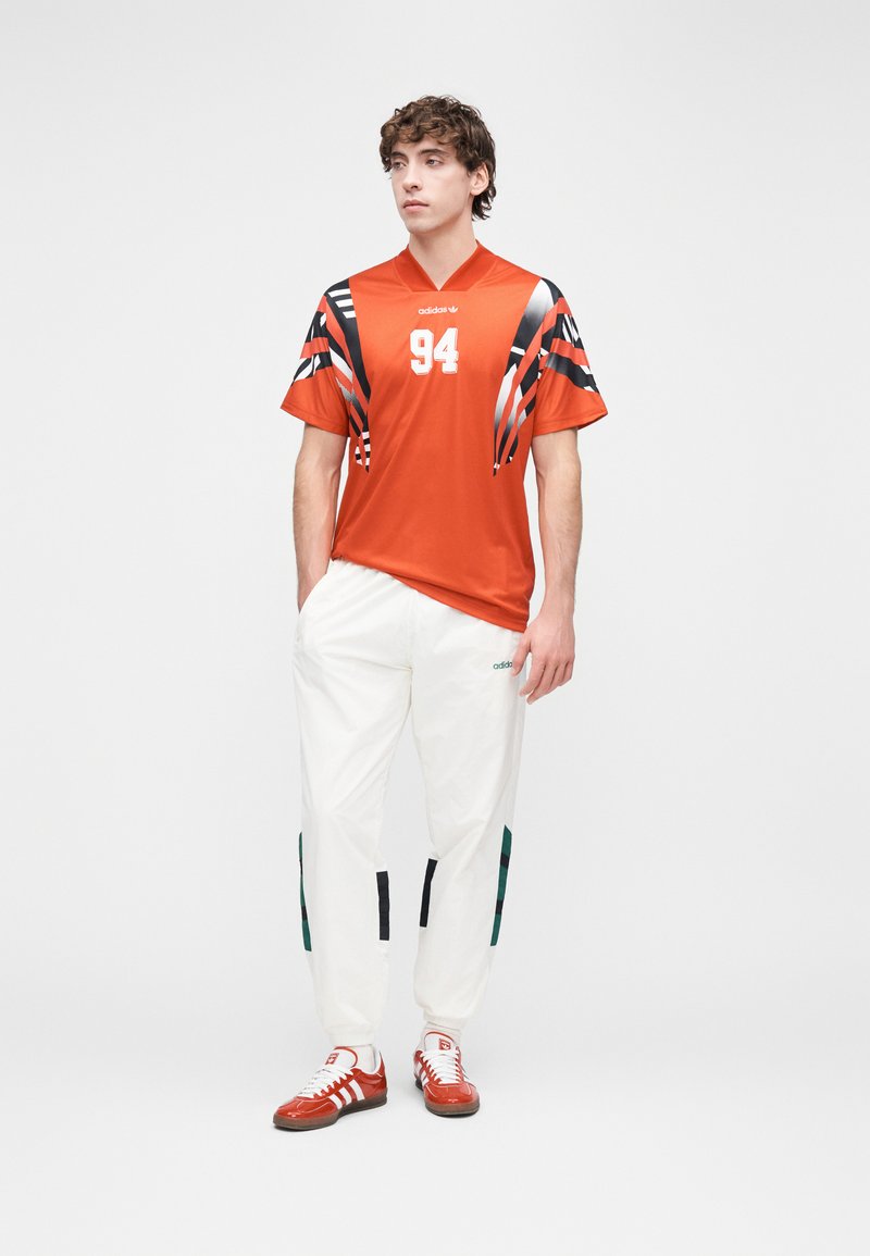Maillot de sport orange avec des accents rayés noirs et blancs et le numéro 94. Associé à un pantalon blanc avec des détails noirs et verts, et des chaussures rouges.