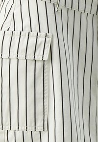 Pantalons clairs avec des rayures verticales noires, dotés d'une poche latérale, fabriqués en tissu coton avec une texture lisse et des détails cousus.