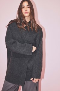 Mladá žena s dlhými hnedými vlasmi, ktorá má na sebe veľký tmavosivý pletený kardigán a voľné sivé nohavice, stojí proti svetloružovému pozadiu.