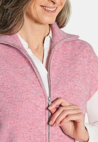 Helles rosa ärmelloses Oberteil mit Reißverschluss, gerippter Textur und hohem Kragen. Über einem weißen Hemd getragen, das eine glatte Oberfläche zeigt.