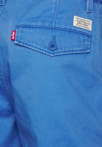 Tela de mezclilla azul claro con un bolsillo trasero que presenta un botón azul y una etiqueta blanca, mostrando el nombre de la marca y los detalles del logotipo.