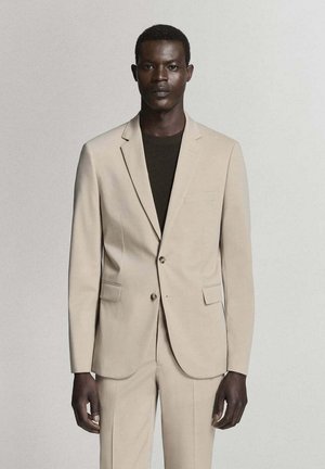 Hombre de piel oscura lleva una chaqueta y pantalones de traje beige sobre una camisa negra, de pie frente a un fondo liso y claro, mirando hacia adelante.