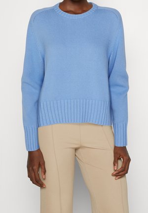Persoon die een lichtblauwe gebreide trui draagt met geribde manchetten en zoom, gecombineerd met beige aansluitende broek tegen een effen achtergrond.