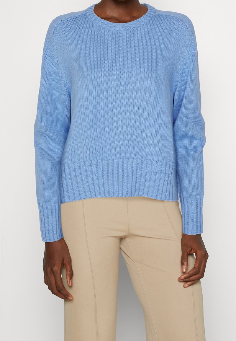 Personne portant un pull en maille bleu clair avec poignets et ourlet côtelés, associé à un pantalon beige ajusté sur un fond uni.
