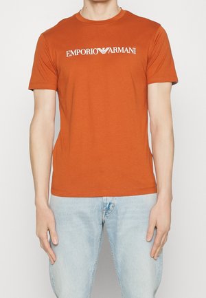 Homme portant un t-shirt orange Emporio Armani avec un texte de logo blanc et un jean bleu clair, debout devant un fond blanc uni.