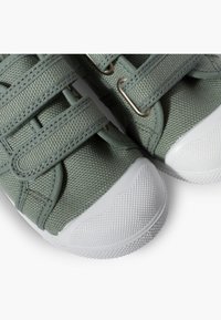 Sneakers in tessuto verde con strappi in Velcro, superficie testurizzata e suole in gomma bianca. Tra i dettagli notevoli ci sono cuciture grigie e accenti in metallo.