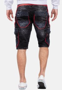 Zwarte denim cargo shorts met rode en blauwe accenten, meerdere zakken en een versleten textuur. De achterkant toont ontwerpdétails en stiksels.