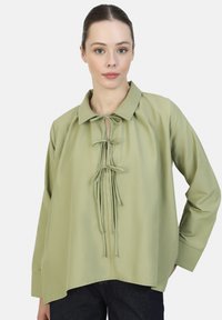 Blusa verde claro de mangas largas con cuello, que presenta un detalle de lazo en la parte frontal y una textura suave y lisa. Corte holgado con dobladillo redondeado.