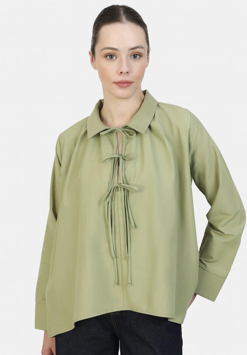 Blusa verde claro de mangas largas con cuello, que presenta un detalle de lazo en la parte frontal y una textura suave y lisa. Corte holgado con dobladillo redondeado.