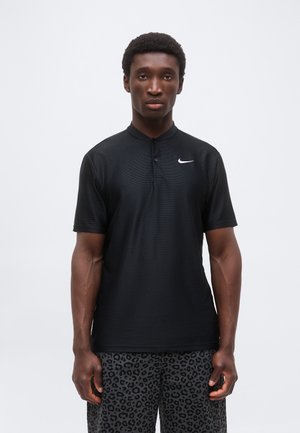 Junger Mann trägt ein schwarzes Nike Kurzarm-Poloshirt und graue Shorts mit Leopardenmuster, steht vor einem schlichten hellen Hintergrund.