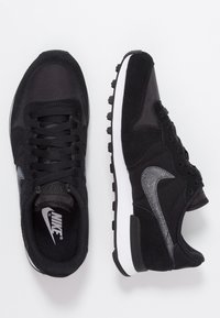 Black athletic shoes featuring mesh and suede materials, a white midsole, and a textured black outsole. Silver Nike swoosh accents.