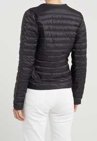 Doudoune noire avec un motif matelassé horizontal, matériau léger, design ajusté et ourlet côtelé. Portée sur un pantalon blanc.