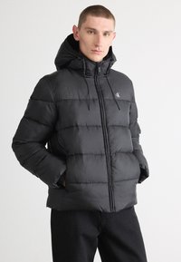 Zwarte gewatteerde jas met een capuchon, voorzien van een ritssluiting, trekkoordaccenten en een logo op de borst. Gladde textuur met horizontale stiksels.