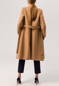 Abrigo beige con cintura ceñida, mangas largas y silueta recta, combinado con pantalones cortos azul marino y tacones oscuros.