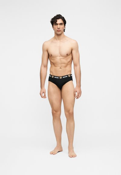 Μυώδης άνδρας όρθιος ξυπόλυτος, φοράει μαύρα εσώρουχα με σχέδιο πρόσωπο αρκούδας και λογότυπο "Moschino" στη μέση, σε απλό λευκό φόντο.