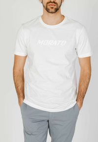 Camiseta de algodón blanca de manga corta y escote redondo. Presenta un logo tonal "MORATO" en el pecho. Combinada con pantalones gris claro.