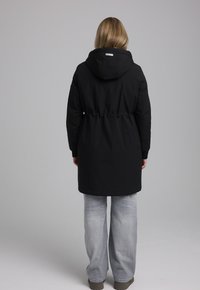 Zwarte gewatteerde parka met een strakke taille, met een gladde textuur en minimalistisch ontwerp. Gecombineerd met lichtgrijze wijde spijkerbroeken.