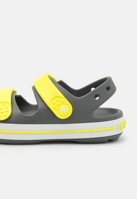 Harmaat sandaalit, joissa on keltainen hihna, valmistettu kumista. Sisältää reikiä ilmanvaihtoa varten, tekstuuripinnan ja valkoisen pohjan keltaisella yksityiskohdalla.