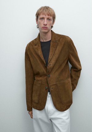 Brown suede blazer with a notched lapel, single button closure, two front pockets, and a relaxed fit. Worn over a black shirt and white pants.