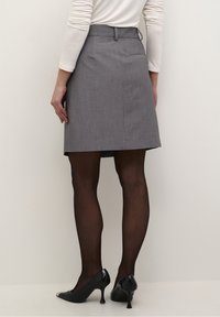 Femme portant une jupe grise mi-longue, des collants noirs transparents, des talons hauts noirs et un haut blanc à manches longues, debout devant un fond uni.