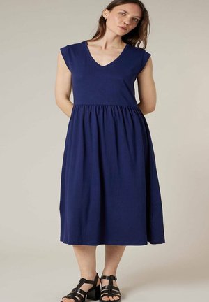 Femme debout portant une robe midi bleu marine sans manches avec une taille froncée et des sandales noires à talons bloc avec brides.