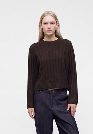 Jeune femme aux cheveux blonds et raides portant un pull côtelé marron foncé et un pantalon bleu foncé, debout devant un fond clair uni.