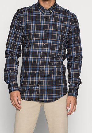 Man draagt een overhemd met lange mouwen met een blauw, zwart en beige ruitpatroon en een beige broek, staand tegen een effen achtergrond.