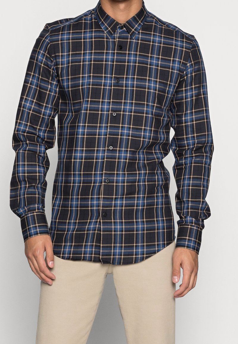 Homme portant une chemise à manches longues à boutons avec un motif à carreaux bleu, noir et beige et un pantalon beige, debout devant un fond uni.