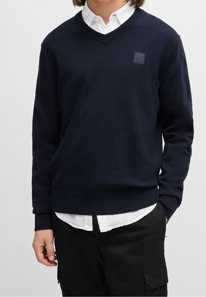 Person iført en mørkeblå V-hals sweater over en hvid knap-på-skjorte og sorte bukser med en hånd i lommen.