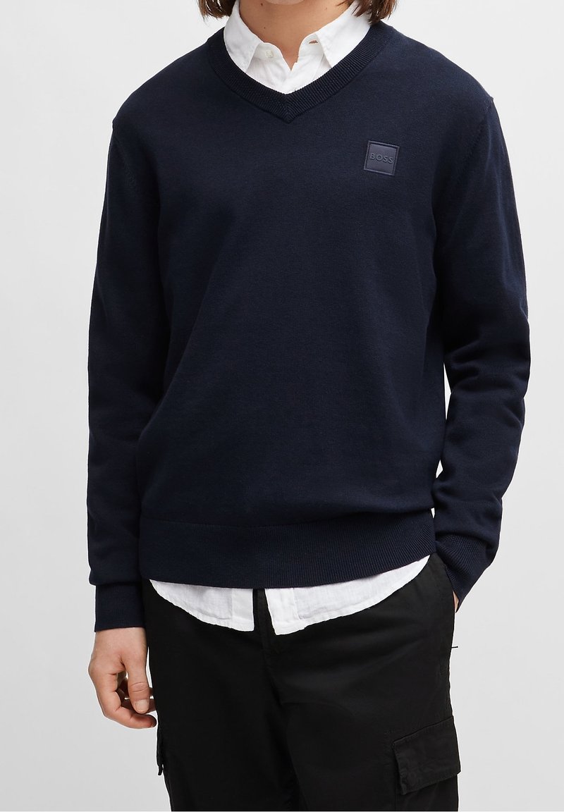 Personne portant un pull bleu marine à col en V par-dessus une chemise blanche boutonnée et un pantalon noir, la main dans la poche.