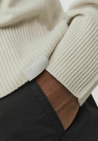 Jasno beżowy sweter dzianinowy o prążkowanej teksturze, z małą metką na mankiecie. Czarny materiał widoczny przy nadgarstku.