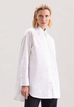 Femme aux cheveux blonds courts portant une chemise blanche oversize boutonnée et un pantalon noir, debout devant un fond clair uni.