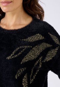 Schwarzer, flauschiger Pullover mit einem strukturierten Goldblatt-Design auf der Vorderseite. Der Stoff hat ein weiches, plüschiges Aussehen und einen weiten Ausschnitt.