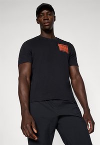 Zwarte katoenen t-shirt met korte mouwen, voorzien van een rode abstracte design op de linkerkant van het borstzakje. Model draagt zwarte broek.