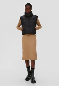 Zwarte puffer vest met een hoge kraag, voorknopsluiting en zijzakken, gedragen over een lichtbruine gebreide jurk tot de knie en zwarte laarzen.