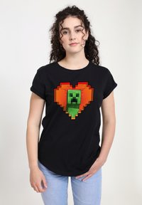 Černé bavlněné tričko s pixelovaným zeleným Creeperem uvnitř oranžového srdce. Krátké rukávy, volný střih a kulatý výstřih.