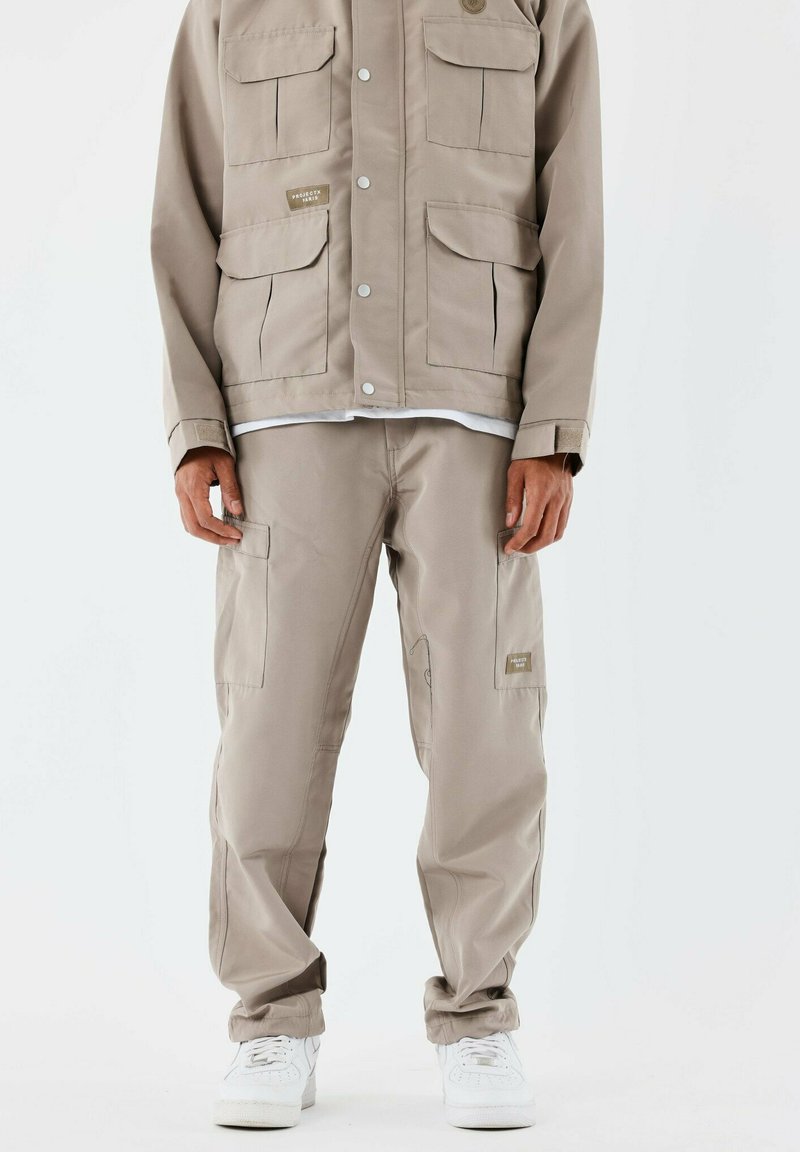 Chaqueta utilitaria beige con múltiples bolsillos, cierres a presión y un par de pantalones cargo beige a juego, que presentan bolsillos laterales y puños elásticos.